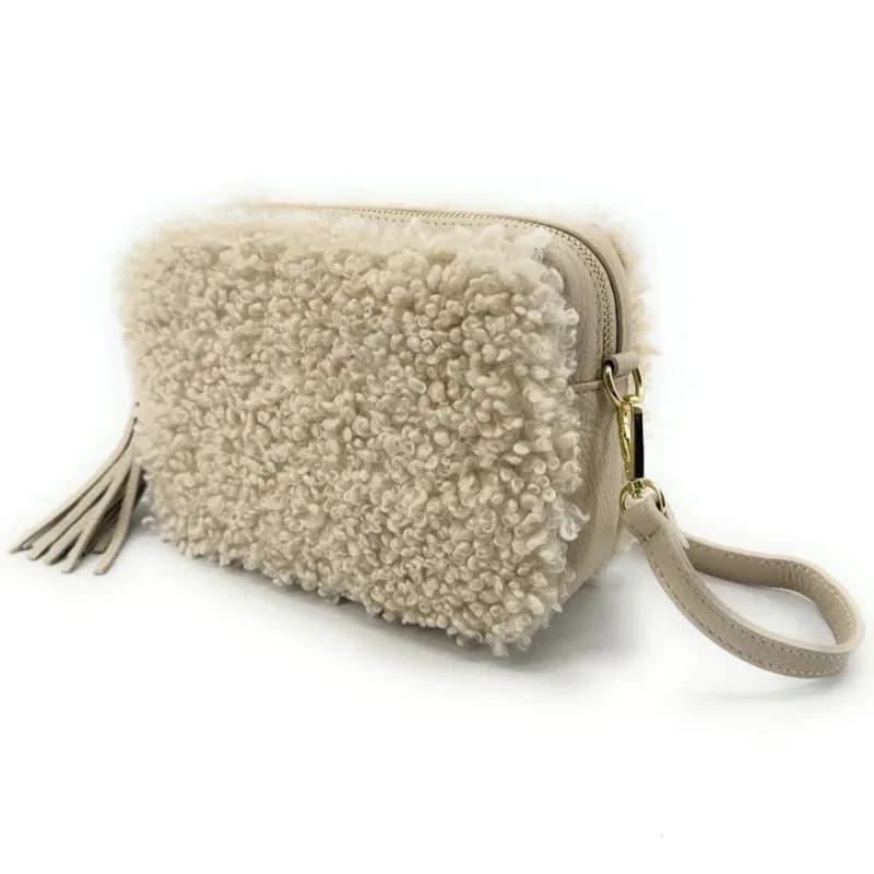 Sac rectangulaire à texture bouclette crème avec pompon, fermeture zippée dorée et dragonne beige, vu de trois quarts sur fond blanc.